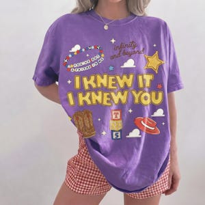 May include: A purple short-sleeve t-shirt featuring yellow text that reads I KNEW IT I KNEW YOU. The graphic includes a beaded bracelet with the text YOU'VE GOT A FRIEND IN ME, a sheriff star, cowboy boots, alphabet blocks, and a red hat.