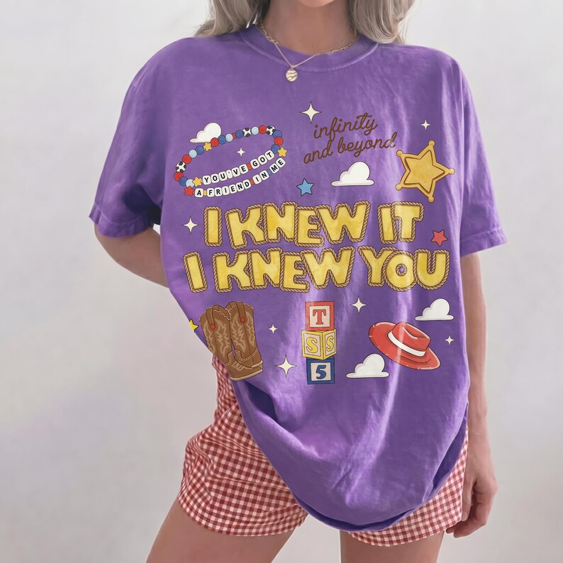 May include: A purple short-sleeve t-shirt featuring yellow text that reads I KNEW IT I KNEW YOU. The graphic includes a beaded bracelet with the text YOU'VE GOT A FRIEND IN ME, a sheriff star, cowboy boots, alphabet blocks, and a red hat.