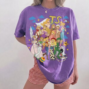 May include: A purple short-sleeve t-shirt featuring a graphic print of multiple blonde characters in various outfits, including a cowgirl, a space ranger, and a sheriff, with the text TS, a rocket, stars, clouds, and a camera.