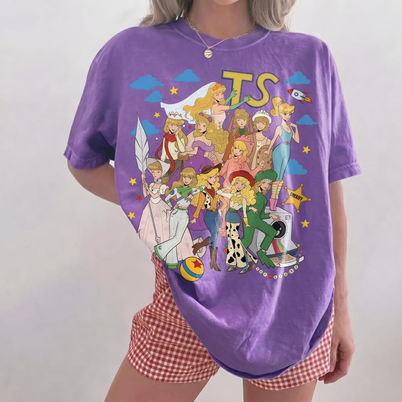 May include: A purple short-sleeve t-shirt featuring a graphic print of multiple blonde characters in various outfits, including a cowgirl, a space ranger, and a sheriff, with the text TS, a rocket, stars, clouds, and a camera.