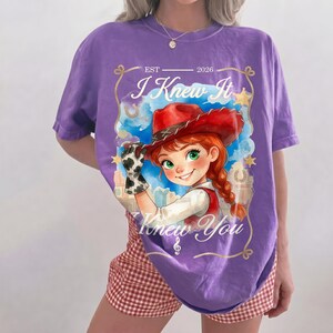 May include: A purple t-shirt featuring a graphic of a cartoon character with red hair in braids wearing a red cowboy hat and a cow-print glove. The text reads EST 2026, I Knew It, and Knew You, framed by a gold rope border with stars and horseshoes.