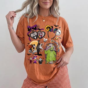 K&ouml;nnte beinhalten: Orangefarbenes T-Shirt mit einem farbenfrohen Grafikdesign mit Figuren aus "The Nightmare Before Christmas". Das Design umfasst Jack Skellington, Sally und Oogie Boogie. Das Shirt wird mit rot-wei&szlig; karierten Shorts kombiniert.