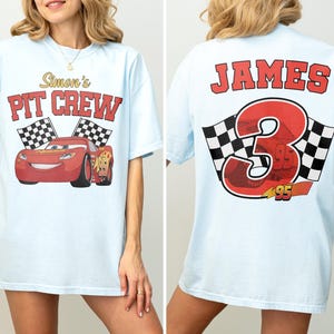 Op de afbeelding: Lichtblauw T-shirt met een rode raceauto-afbeelding en de tekst "Simon's PIT CREW" op de voorkant. De achterkant van het shirt toont de naam "JAMES" boven een groot rood nummer "3" met geblokte vlaggen en het nummer "95".