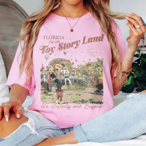 May include: A light pink t-shirt featuring a colorful graphic of Toy Story Land in Florida. The design includes Mickey Mouse, Woody, Buzz Lightyear, and other characters, with the text "Florida Est. 2018 Toy Story Land To Infinity and Beyond."