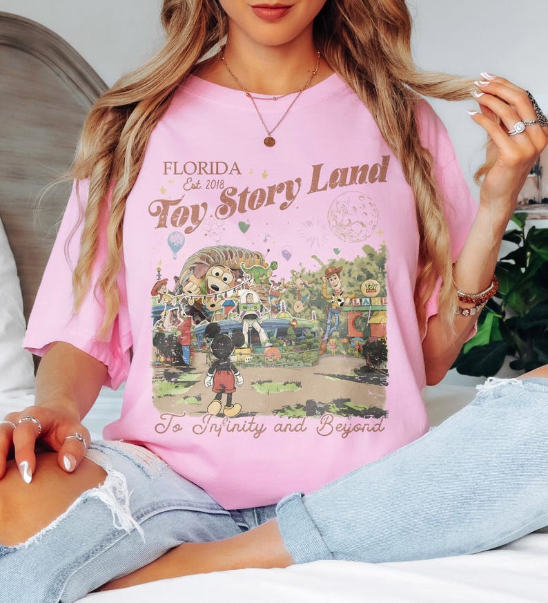 May include: A light pink t-shirt featuring a colorful graphic of Toy Story Land in Florida. The design includes Mickey Mouse, Woody, Buzz Lightyear, and other characters, with the text "Florida Est. 2018 Toy Story Land To Infinity and Beyond."
