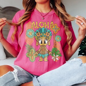 May include: A vibrant pink t-shirt with a tropical-themed graphic. The design features the word "ALOHA" above a cartoon duck in a Hawaiian headdress, with floral and geometric elements in teal, orange, and green.