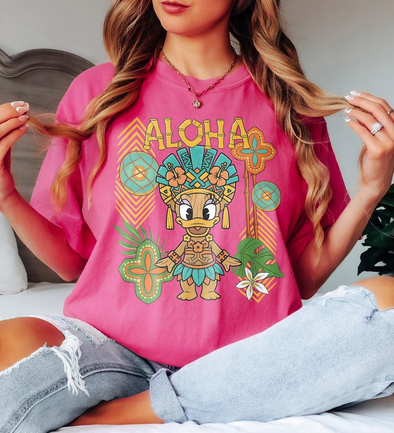 May include: A vibrant pink t-shirt with a tropical-themed graphic. The design features the word "ALOHA" above a cartoon duck in a Hawaiian headdress, with floral and geometric elements in teal, orange, and green.