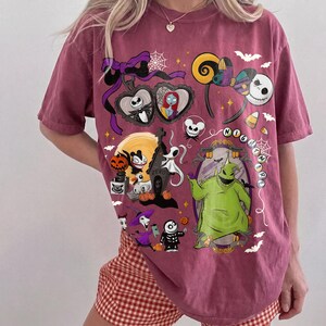 K&ouml;nnte beinhalten: Ein weinrotes T-Shirt mit einem Design mit Figuren aus "The Nightmare Before Christmas". Die Grafik zeigt Jack Skellington, Sally, Zero und Oogie Boogie sowie andere Halloween-Motive. Das Shirt hat einen Rundhalsausschnitt und kurze &Auml;rmel.