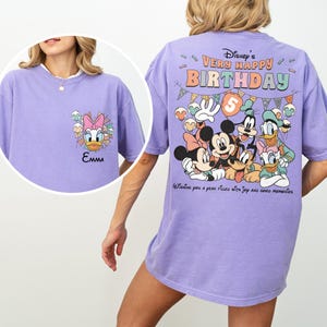 Puede incluir: Una camiseta de color lila claro con personajes de Disney. La parte delantera presenta a Daisy Duck y el nombre "Emma". La parte trasera muestra "Disney's Very Happy Birthday" con Mickey, Minnie, Donald, Goofy y Pluto. La camiseta tiene un corte holgado.