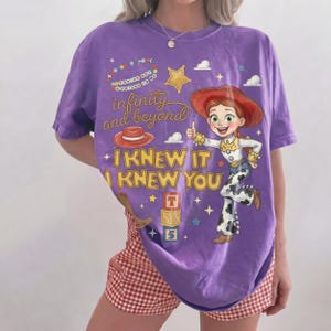 May include: A purple short-sleeve t-shirt featuring a graphic of a cowgirl character, a red hat, and stacked alphabet blocks. The text reads "YOU'VE GOT A FRIEND IN ME", "infinity and beyond", and "I KNEW IT I KNEW YOU" in yellow and white.