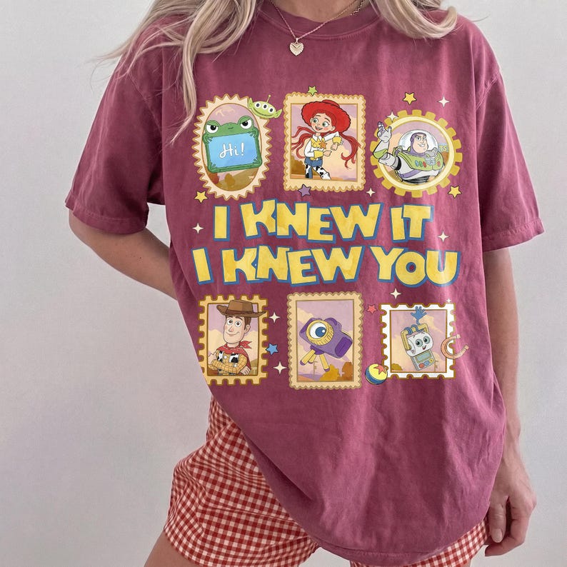 May include: A dusty rose short-sleeve t-shirt featuring a graphic print of Toy Story characters in postage stamp frames. The text reads I KNEW IT I KNEW YOU in yellow letters. The design includes images of Woody, Jessie, Buzz Lightyear, and others.