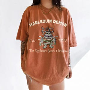 Op de afbeelding: Verbrand oranje t-shirt met een afbeelding van een monster en de tekst "HARLEQUIN DEMON". Onder het monster staat de tekst "Est. 1993 The Nightmare Before Christmas". Het shirt heeft korte mouwen en een losse pasvorm.