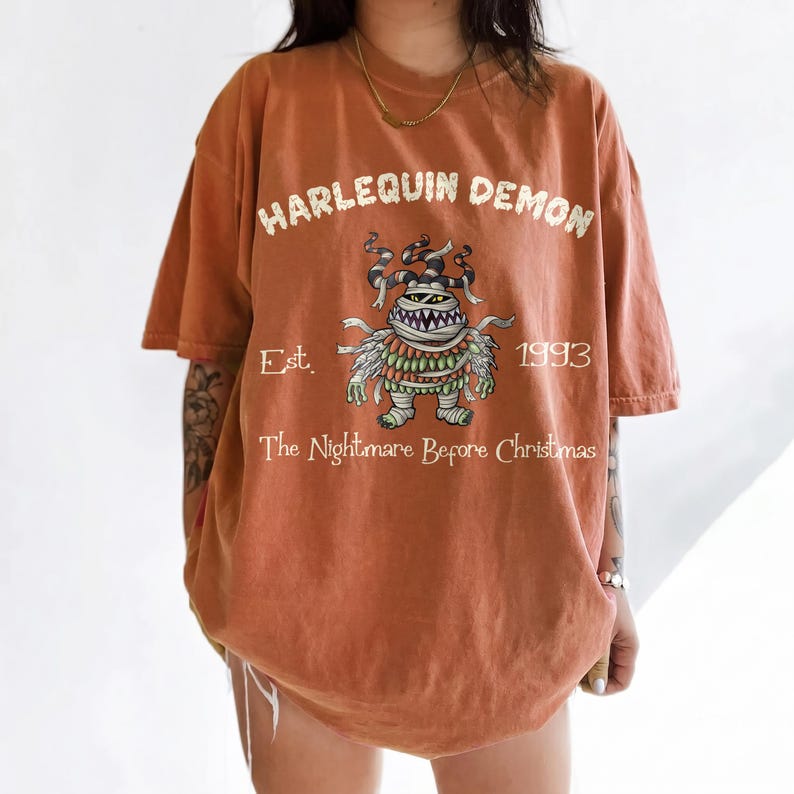 Op de afbeelding: Verbrand oranje t-shirt met een afbeelding van een monster en de tekst "HARLEQUIN DEMON". Onder het monster staat de tekst "Est. 1993 The Nightmare Before Christmas". Het shirt heeft korte mouwen en een losse pasvorm.