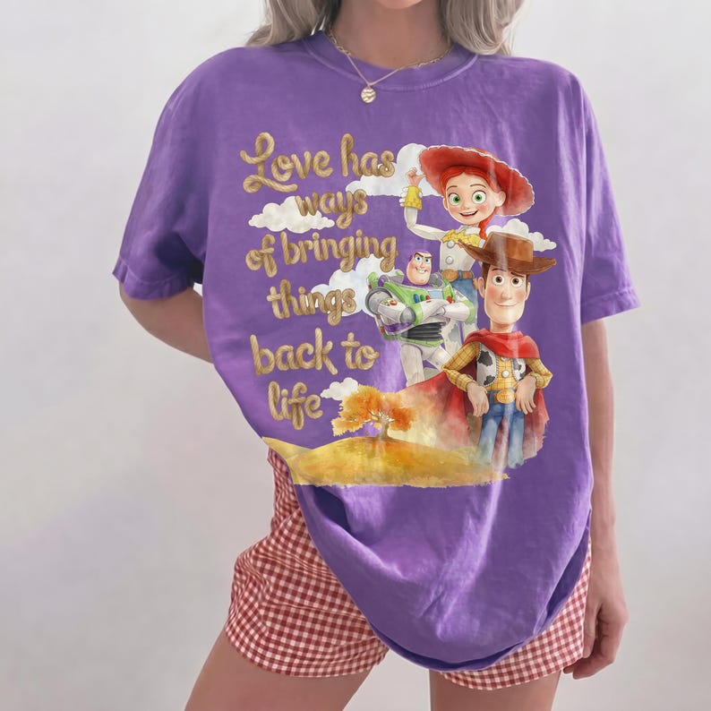 Pu&ograve; includere: Una t-shirt viola a maniche corte con una grafica di Woody, Buzz Lightyear e Jessie di Toy Story. Il testo "Love has ways of bringing things back to life" &egrave; stampato in caratteri dorati sopra un paesaggio giallo con un albero.