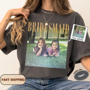 Könnte beinhalten: Ein anthrazitfarbenes T-Shirt mit dem Wort BRIDESMAID in goldener Schrift über einem aufgedruckten Foto von zwei jungen Kindern, die auf Gras liegen. Das T-Shirt enthält eine kleine grafische Einblendung mit dem Text Your photo.