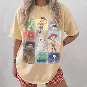 K&ouml;nnte beinhalten: Ein blassgelbes Kurzarm-T-Shirt mit einem Raster aus neun farbenfrohen Illustrationen animierter Figuren aus der Toy Story-Filmreihe, darunter Woody, Buzz Lightyear, Jessie, Rex, Slinky Dog, Mr. und Mrs. Potato Head, Bullseye und Forky.