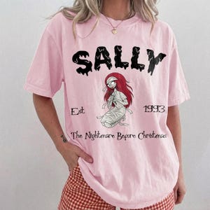 Op de afbeelding: Lichtroze T-shirt met de naam "SALLY" in zwarte druipende letters boven een illustratie van Sally uit "The Nightmare Before Christmas". Het ontwerp bevat de tekst "Est 1993" en "The Nightmare Before Christmas."