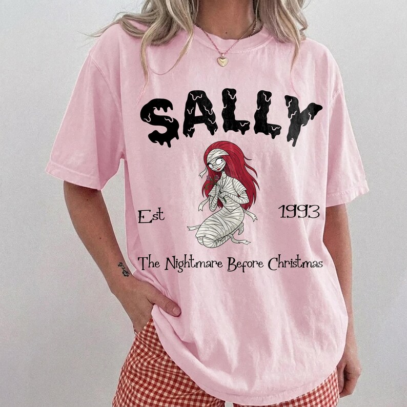 Op de afbeelding: Lichtroze T-shirt met de naam "SALLY" in zwarte druipende letters boven een illustratie van Sally uit "The Nightmare Before Christmas". Het ontwerp bevat de tekst "Est 1993" en "The Nightmare Before Christmas."