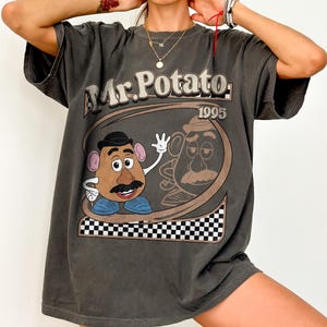 May include: Dark gray t-shirt featuring a Mr. Potato Head graphic. The design includes the text "Mr. Potato 1995" in a brown and white color scheme. The shirt has a vintage, worn look.