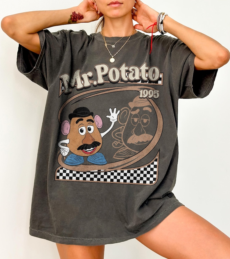 May include: Dark gray t-shirt featuring a Mr. Potato Head graphic. The design includes the text "Mr. Potato 1995" in a brown and white color scheme. The shirt has a vintage, worn look.