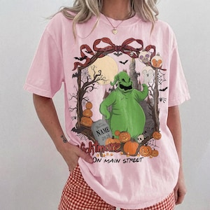 May include: A light pink t-shirt featuring a graphic of a green character, pumpkins, a tombstone with the text NAME 2026, and a castle silhouette. The design includes a red bow, bats, spiderwebs, and the text Nightmare ON MAIN STREET.