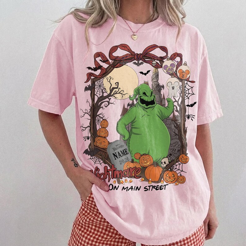 May include: A light pink t-shirt featuring a graphic of a green character, pumpkins, a tombstone with the text NAME 2026, and a castle silhouette. The design includes a red bow, bats, spiderwebs, and the text Nightmare ON MAIN STREET.