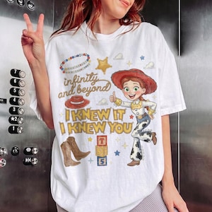 May include: A white t-shirt featuring a graphic print of a cowgirl character, a red cowboy hat, brown cowboy boots, and colorful alphabet blocks. The text reads "infinity and beyond" and "I KNEW IT I KNEW YOU" along with a beaded necklace design.