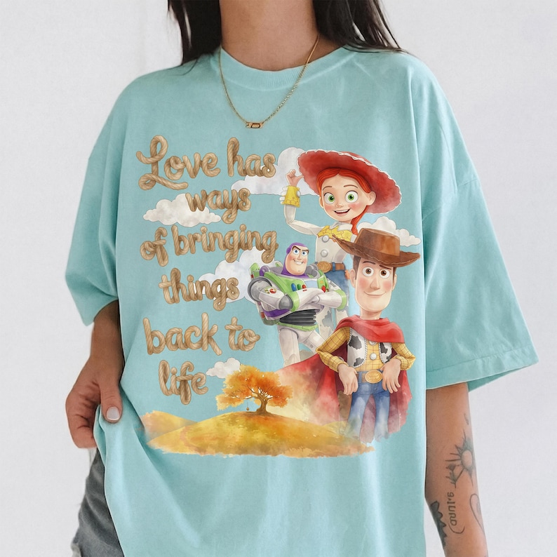 Pu&ograve; includere: Una t-shirt azzurra con una stampa grafica di Woody, Buzz Lightyear e Jessie di Toy Story. Il design include un albero dorato su una collina e il testo "Love has ways of bringing things back to life" in un carattere corsivo color marrone dorato.