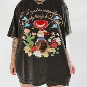 May include: A black t-shirt featuring a graphic of a cowgirl in a red hat riding a horse, surrounded by cacti and colorful flowers. The text reads, Love has ways of bringing things back to life, accompanied by small blue clouds and musical notes.