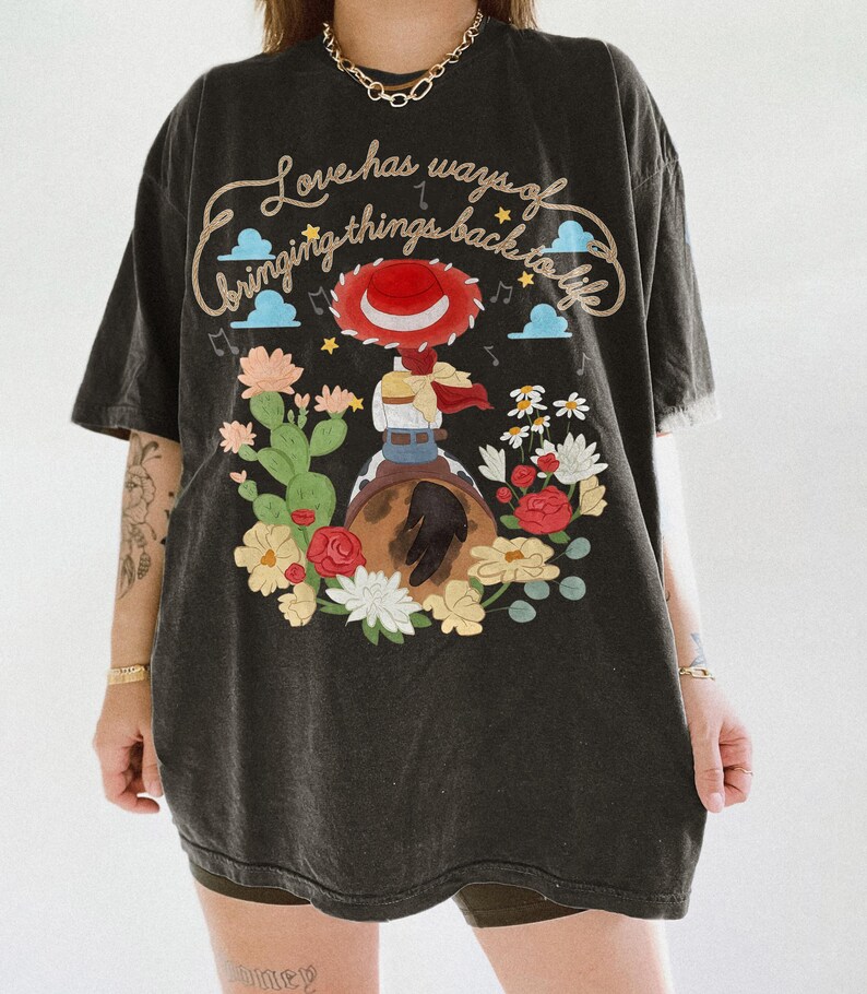 May include: A black t-shirt featuring a graphic of a cowgirl in a red hat riding a horse, surrounded by cacti and colorful flowers. The text reads, Love has ways of bringing things back to life, accompanied by small blue clouds and musical notes.