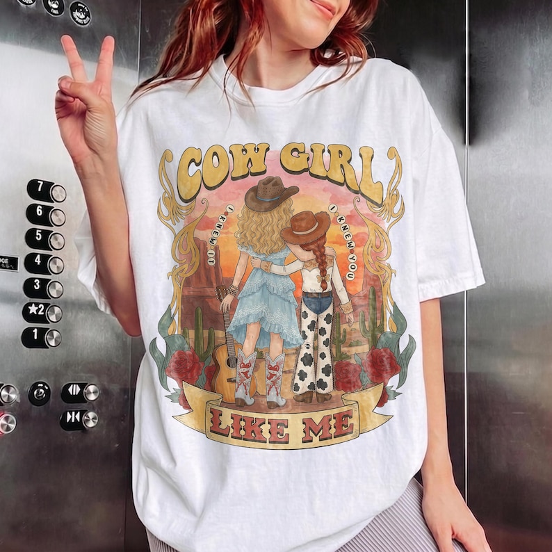 May include: A white t-shirt featuring a graphic print of two figures in cowboy hats standing in a desert landscape with cacti and red roses. The text COW GIRL LIKE ME is printed in large letters. One figure wears a blue dress and the other cow print pants.