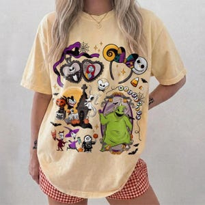K&ouml;nnte beinhalten: Hellgelbes T-Shirt mit einem farbenfrohen Grafikdesign mit Charakteren und Elementen aus "The Nightmare Before Christmas". Das Design umfasst Jack Skellington, Sally, Oogie Boogie und andere ikonische Bilder. Das Shirt hat eine lockere Passform.