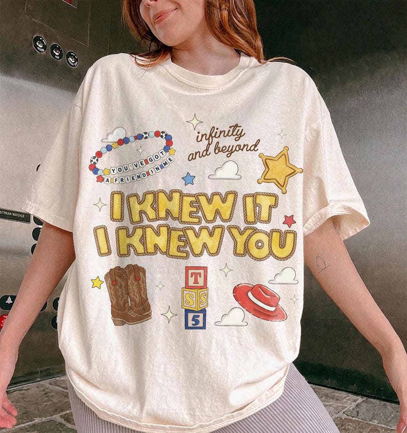 May include: A cream-colored short-sleeve t-shirt featuring a graphic print with the text "I KNEW IT I KNEW YOU" in yellow, "infinity and beyond" in black, and "YOU'VE GOT A FRIEND IN ME" on a beaded bracelet illustration, plus cowboy boots, a hat, and blocks.