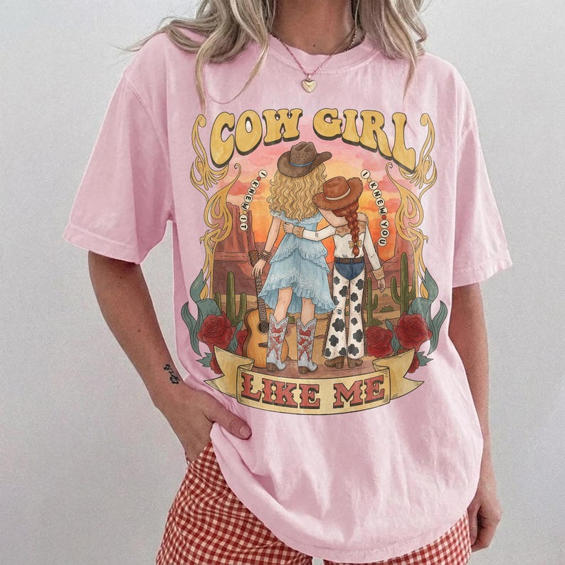 May include: A light pink short-sleeve t-shirt featuring a graphic print of two figures in cowboy hats standing in a desert landscape with cacti and red roses. The text "COW GIRL" appears at the top and "LIKE ME" on a banner at the bottom.