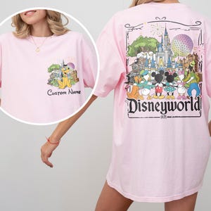 May include: Light pink t-shirt featuring a Disney World graphic on the back and a smaller graphic on the front. The back graphic includes the text "Disneyworld 1971" and various Disney characters. The front graphic has Pluto and the text "Custom Name".