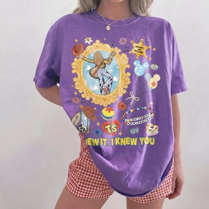 May include: A purple short-sleeve t-shirt featuring a graphic print of a person with a guitar inside a gold frame, a sheriff star with the text INFINITY AND BEYOND, a ball, a heart with TS, and the text YOU'VE GOT A FRIEND IN ME and KNEW YOU.
