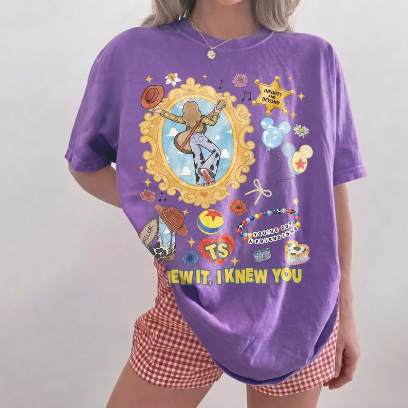May include: A purple short-sleeve t-shirt featuring a graphic print of a person with a guitar inside a gold frame, a sheriff star with the text INFINITY AND BEYOND, a ball, a heart with TS, and the text YOU'VE GOT A FRIEND IN ME and KNEW YOU.