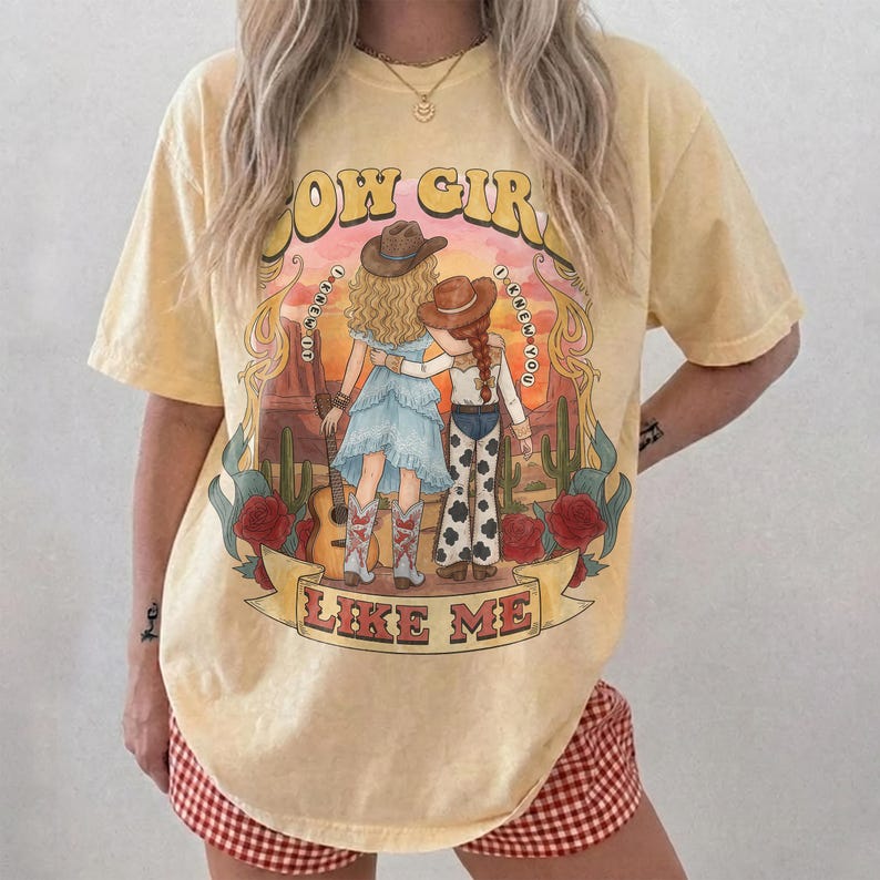 May include: A pale yellow t-shirt featuring a graphic print of two figures in cowboy hats standing in a desert landscape with cacti and red roses. The text reads COW GIRLS I KNEW DO LIKE ME. The design includes a guitar and western motifs.