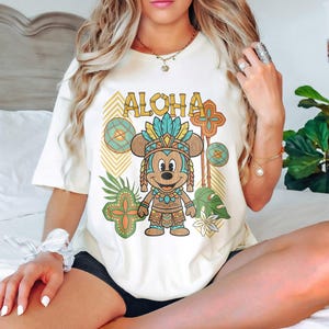 May include: Cream-coloured t-shirt featuring a cartoon Mickey Mouse in tribal attire with the word "ALOHA" in a gold font. The design includes tropical leaves and geometric patterns in shades of green, orange, and blue. The shirt is a relaxed fit.