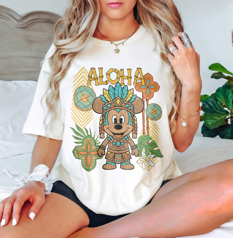 May include: Cream-coloured t-shirt featuring a cartoon Mickey Mouse in tribal attire with the word "ALOHA" in a gold font. The design includes tropical leaves and geometric patterns in shades of green, orange, and blue. The shirt is a relaxed fit.