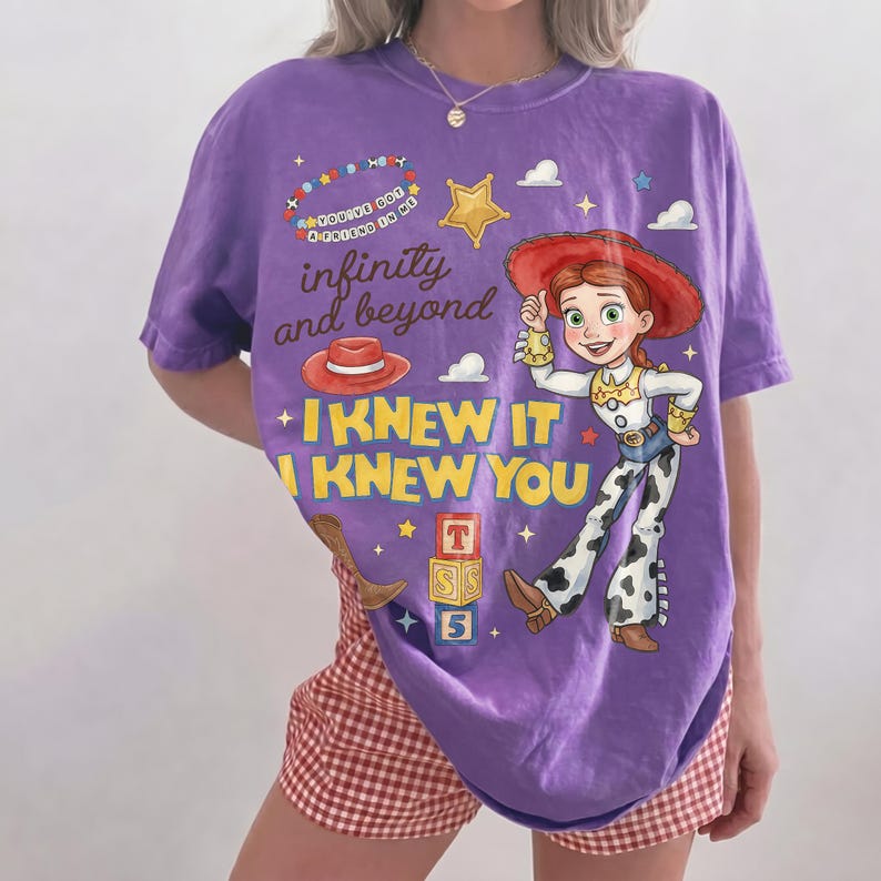 Pode incluir: Uma t-shirt roxa de manga curta com o gr&aacute;fico de uma cowgirl com chap&eacute;u vermelho, camisa amarela e cal&ccedil;as com manchas de vaca. O design inclui o texto "infinity and beyond", "I KNEW IT", "I KNEW YOU", um chap&eacute;u vermelho e blocos.