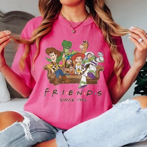 May include: A pink t-shirt featuring a graphic of characters from the Toy Story movie franchise. The characters are arranged on a brown couch, with the word "FRIENDS" above "SINCE 1995" in black text.