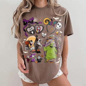 K&ouml;nnte beinhalten: Braunes T-Shirt mit einem farbenfrohen Grafikdesign mit Charakteren und Elementen aus "The Nightmare Before Christmas". Das Design umfasst Jack Skellington, Sally, Oogie Boogie und andere ikonische Bilder. Das Shirt hat einen Rundhalsausschnitt und kurze &Auml;rmel.