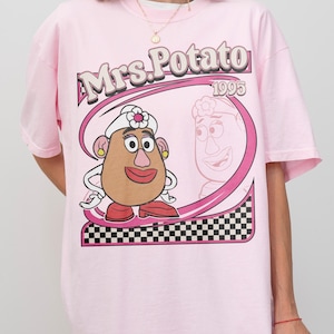 May include: Pink t-shirt featuring a cartoon image of Mrs. Potato Head with the text "Mrs. Potato 1995". The graphic includes a pink oval frame with a black and white checkered pattern at the bottom.