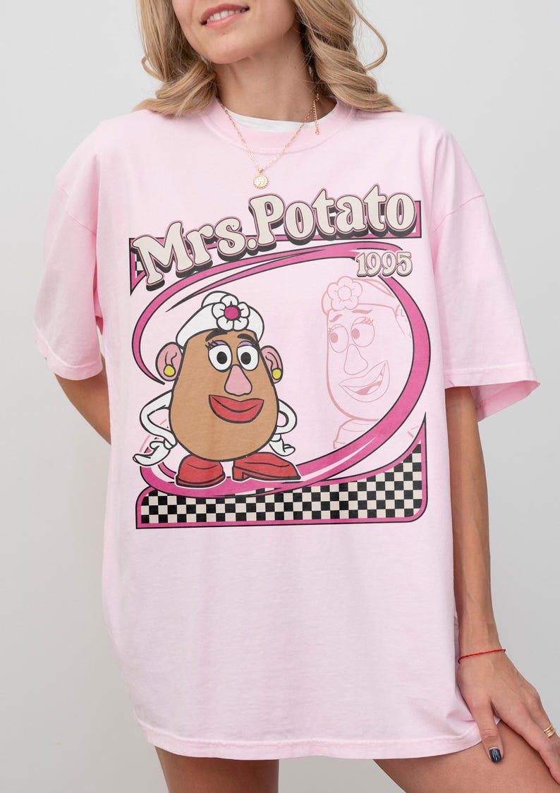May include: Pink t-shirt featuring a cartoon image of Mrs. Potato Head with the text "Mrs. Potato 1995". The graphic includes a pink oval frame with a black and white checkered pattern at the bottom.