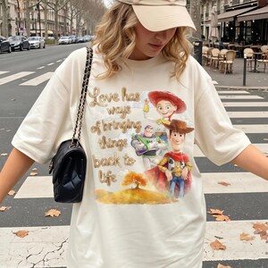 Pu&ograve; includere: Una t-shirt color crema con una stampa grafica di Woody, Buzz Lightyear e Jessie di Toy Story, con il testo "Love has ways of bringing things back to life" sopra un paesaggio in stile acquerello con un albero dorato.
