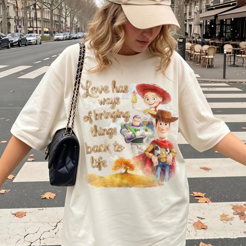 Pu&ograve; includere: Una t-shirt color crema con una stampa grafica di Woody, Buzz Lightyear e Jessie di Toy Story, con il testo "Love has ways of bringing things back to life" sopra un paesaggio in stile acquerello con un albero dorato.