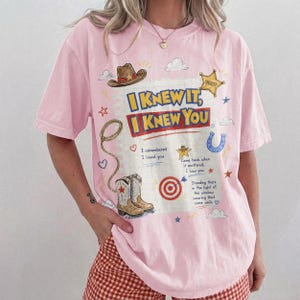 May include: A light pink short-sleeve t-shirt featuring a graphic print of a cowboy hat, a sheriff star, a lasso, and cowboy boots. The text reads: I KNEW IT, I KNEW YOU, I remembered I loved you, Came back when it mattered, I saw you, Standing there in the light of the window wearing that same smile.