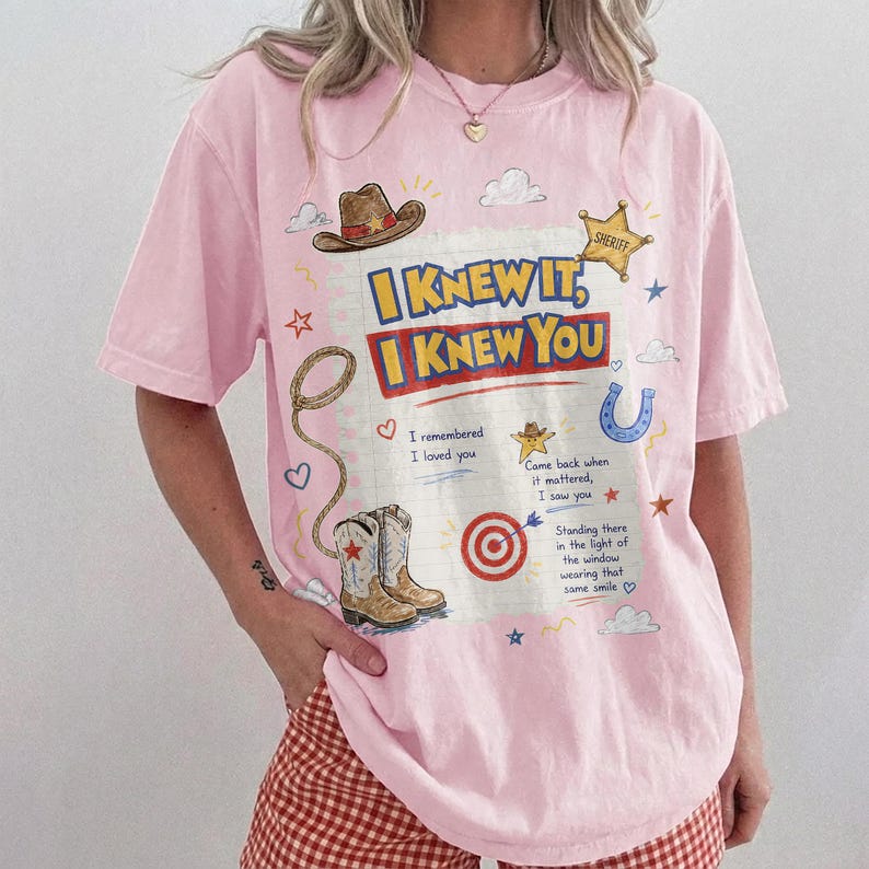 May include: A light pink short-sleeve t-shirt featuring a graphic print of a cowboy hat, a sheriff star, a lasso, and cowboy boots. The text reads: I KNEW IT, I KNEW YOU, I remembered I loved you, Came back when it mattered, I saw you, Standing there in the light of the window wearing that same smile.