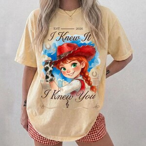 May include: A pale yellow t-shirt featuring a graphic of a cartoon cowgirl with red hair and a red cowboy hat. The text reads "EST 2026", "I Knew It", and "I Knew You". The design includes a blue watercolor splash background and a musical note.